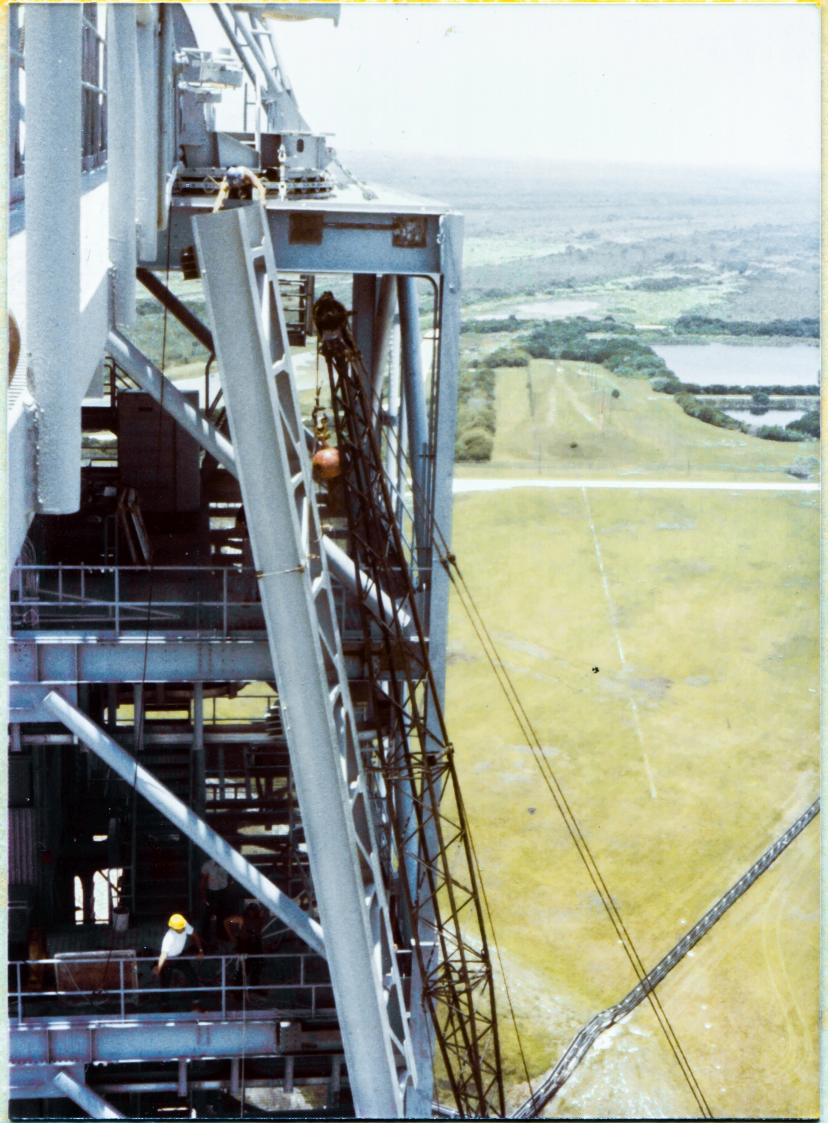 Image 078. Leaning dangerously out over empty space from a kneeling position at Elevation 300'-0”, unharnessed, untethered, Union Ironworker James Dixon barehands the top of the GOX Arm Hinges Support Strongback where it meets the top of the Fixed Service Structure at Space Shuttle Launch Complex 39-B, Kennedy Space Center, Florida, as another, unknown alas, Union Ironworker in a white shirt wearing a yellow hardhat, two levels below at Elevation 260'-0”, gives hand signals to the crane operator, at the controls in his cab, down on the Pad Deck at Elevation 53'-0”. The crane operator has no way to see what's going on far above him with any kind of detail, and relies exclusively on the hand signals he's being given, even as everyone else relies on his skill at swinging, rotating, and nudging forty feet of steel truss weighing well over 10,000 pounds into a final position that has to wind up within roughly one sixteenth of an inch of where it's needed for final attachment to the FSS. Everyone's lives hang in the balance as this work is done, they all know this, and they all trust one another. With their own lives. As they do the work. Photo by James MacLaren.